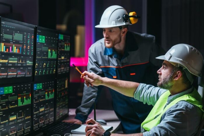Twee operators met helmen en veiligheidsvest analyseren gegevens op meerdere monitoren in een controlekamer